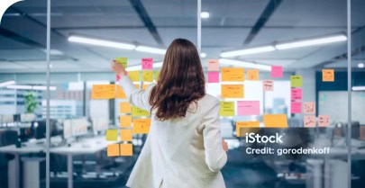Frau in Businesskleidung klebt bunte Post-its an eine Glaswand im modernen Büro.